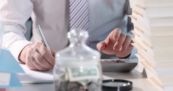 Бізнесмен розраховує на калькулятор і пише на папері перед скляною банкою з грошима крупним планом 4k фільм повільний motio — стокове відео