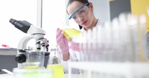 Mujer química examinando líquido amarillo en frasco delante del microscopio en laboratorio 4k película cámara lenta — Vídeos de Stock