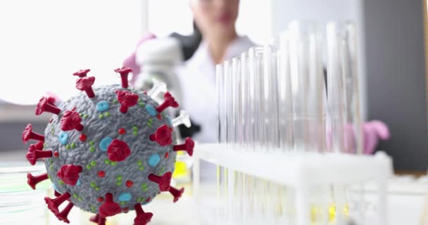 Искусственная модель коронавируса лежит на столе перед химиком-женщиной с микроскопом 4k фильм замедленной съемки — стоковое видео