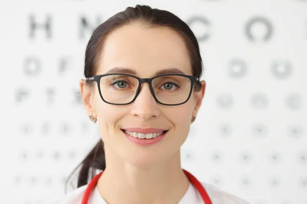 Glimlachende vrouwelijke oogarts met oogtest op achtergrond, vrouwelijke opticien in bril — Stockfoto