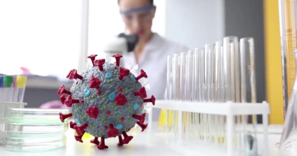 Искусственная модель коронавируса лежит на столе перед ученым, смотрящим в микроскоп 4k фильма замедленной съемки — стоковое видео