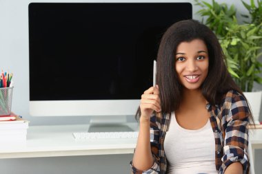 Afro-Amerikalı genç bayan masada bilgisayar aygıtıyla ofiste oturuyor.