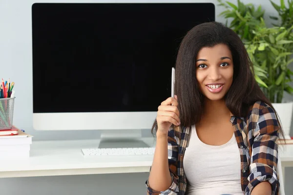 Afro américaine jeune femme assis dans le bureau avec dispositif informatique derrière sur le bureau — Photo