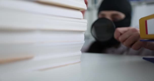 Thief in black balaclava looking into folders with documents using magnifying glass 4k movie slow motion — Stockvideo