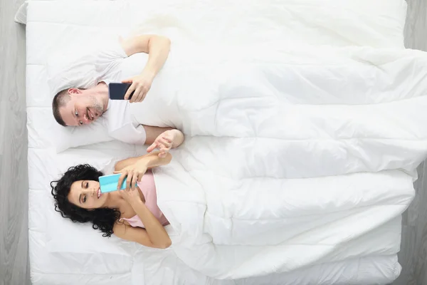 Esposa y marido acostados en la cama mirando las pantallas de los teléfonos desde temprano en la mañana —  Fotos de Stock