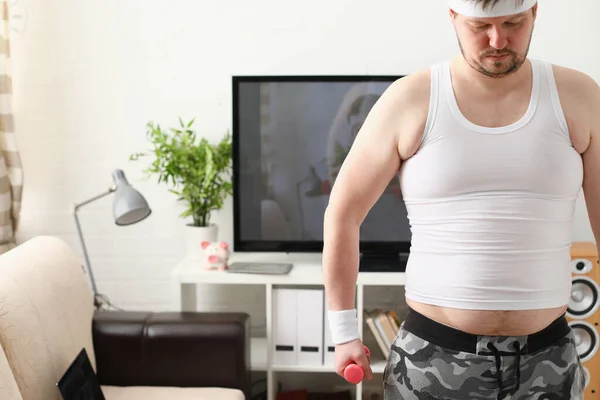 Overweight man is engaged in fitness at home holding dumbbell in hand — Stockfoto
