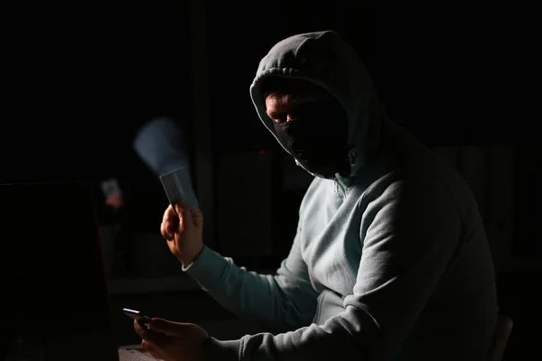 Hacker en chaqueta oscura mirando la pantalla del ordenador portátil en la habitación oscura es la celebración de la tarjeta bancaria —  Fotos de Stock