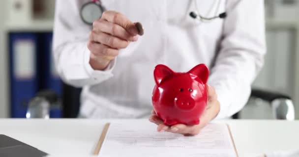 Doctor throws coin into piggy bank closeup — Wideo stockowe