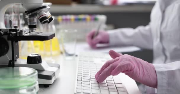 Scientist types on keyboard and takes notes on documents in laboratory — Stockvideo