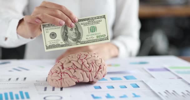 Женская рука кладет стодолларовую купюру в макет человеческого мозга — стоковое видео