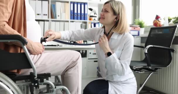 Mujer escucha vientre con estetoscopio a mujer embarazada en silla de ruedas. — Vídeos de Stock