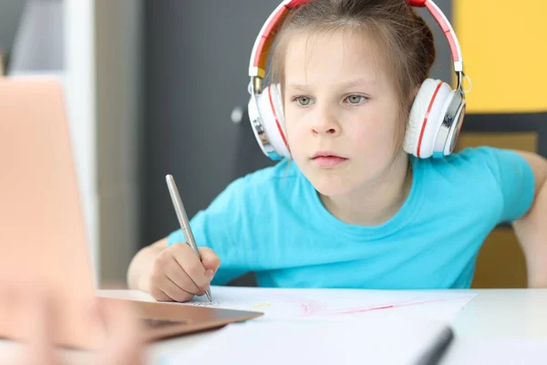 Petite fille écolière dans les écouteurs assis devant un ordinateur portable à la maison — Photo