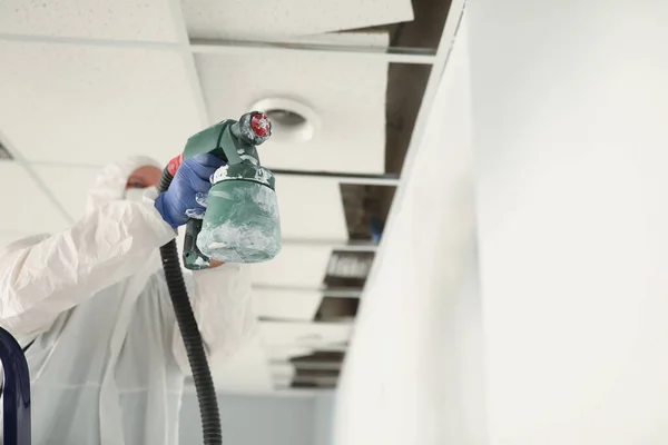 Koruyucu tulumlu erkek inşaat işçisi duvarları sprey silahla beyaza boyuyor. — Stok fotoğraf