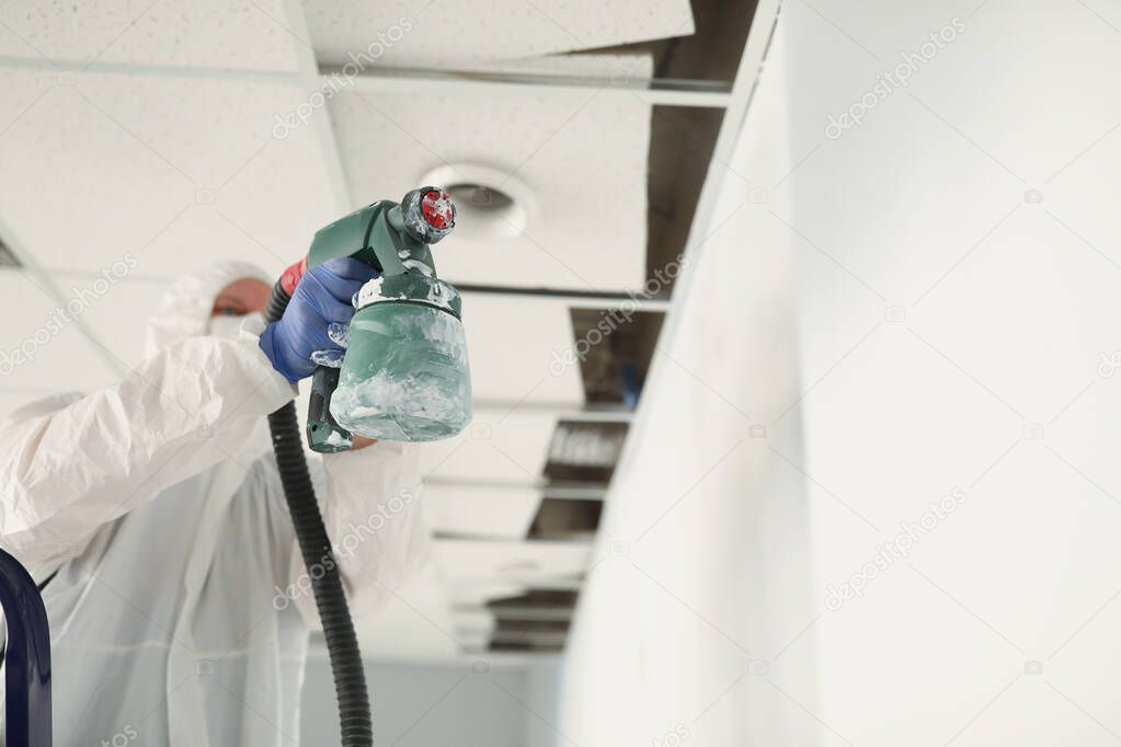 Male construction worker in protective overalls painting wall white with spray gun closeup