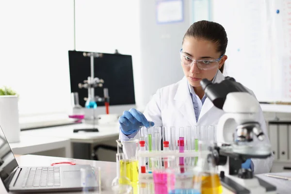Женщина-химик держит пробирку с жидкостью перед микроскопом в лаборатории — стоковое фото