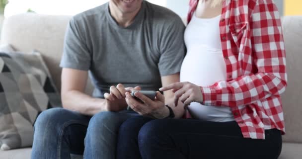 Marido e mulher grávida sentados no sofá e olhando para a tela do telefone celular filme 4k — Vídeo de Stock