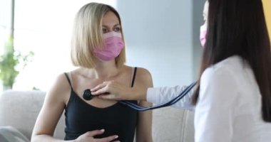 Koruyucu tıbbi maskeli doktor, 4K filminde steteskop kullanan kadın hastayı dinliyor.