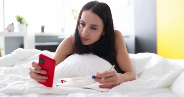 Nieszczęśliwa kobieta patrząc test ciążowy i trzymając smartfona leżącego na łóżku — Wideo stockowe