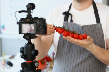 Vlogger şefi olgun domates malzemesi gösteriyor, önlüklü adam kırmızı sebze kaydediyor.