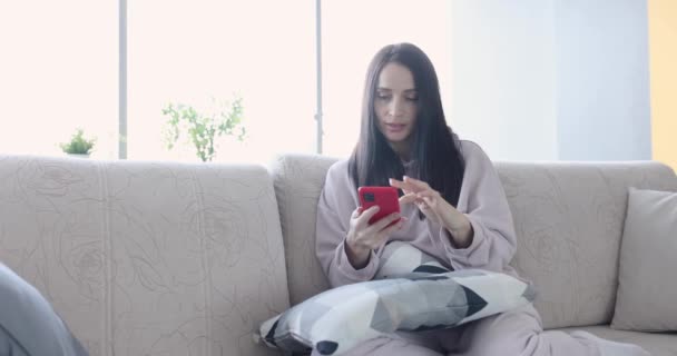 Kobieta siedząca na kanapie z telefonem komórkowym i uśmiechniętym filmem 4k — Wideo stockowe