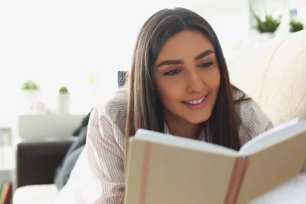 Giovane donna attraente lettura libro sdraiato sul divano a casa, storia interessante — Foto Stock