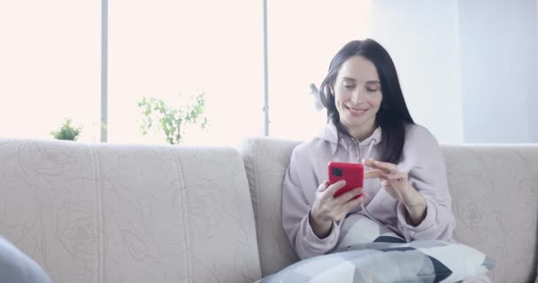 Mujer sonriente mirando en el teléfono inteligente en el sofá — Vídeos de Stock
