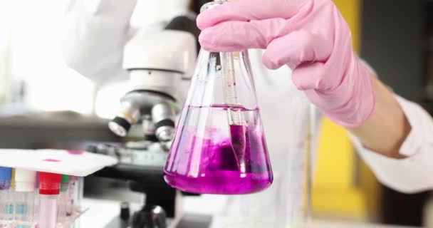 Хімік тримає колбу з прозорою рідиною в руках і кидає в неї фіолетовий розчинник — стокове відео