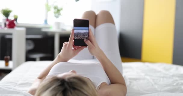 Žena leží na posteli a dívá se na fotky z dovolené na telefonu — Stock video