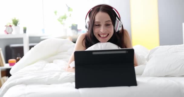 Mulher sorridente em fones de ouvido se comunica por chamada de vídeo enquanto deitado na cama — Vídeo de Stock