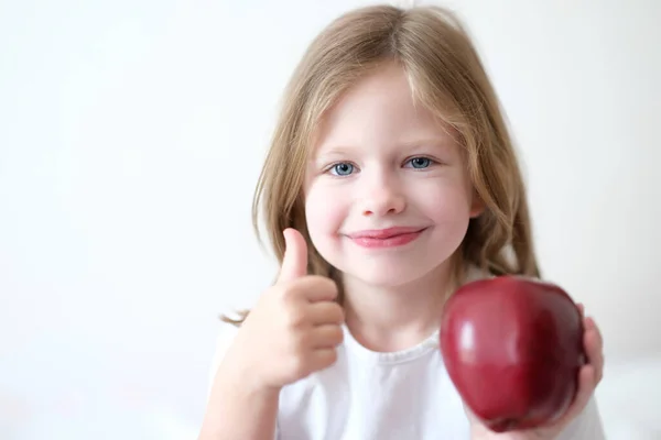 En liten flicka som håller ett rött äpple i händerna och visar tummen upp — Stockfoto