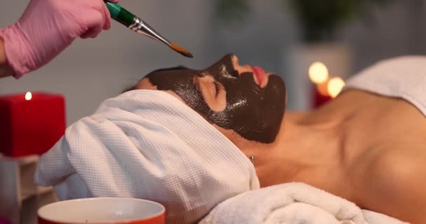 Esteticista aplicando máscara de arcilla oscura en la cara de la mujer — Vídeos de Stock