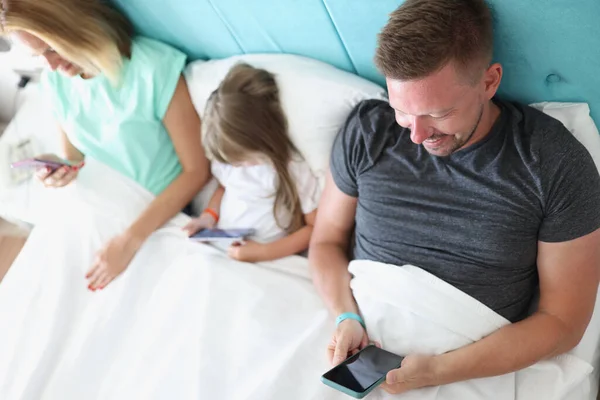 Молода сім'я з дитиною використовує гаджети, лежачи разом у ліжку — стокове фото