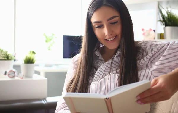 Młoda uśmiechnięta kobieta czyta książkę siedząc na kanapie w domu, ciekawa historia — Zdjęcie stockowe