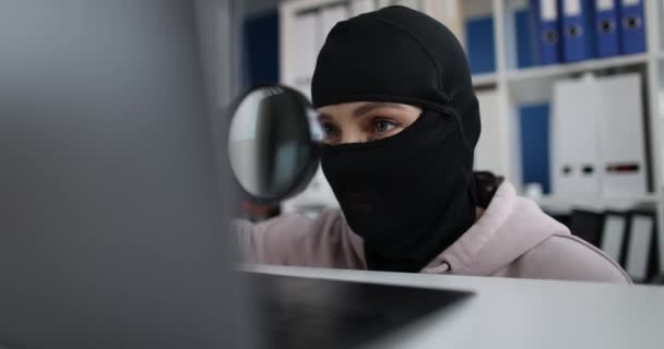 Robber wearing a mask and searching for information using magnifying glass on computer in office — Stockvideo