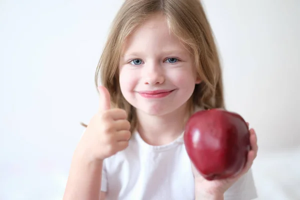 Liten flicka håller rött äpple i händerna och visar tummen upp gest — Stockfoto