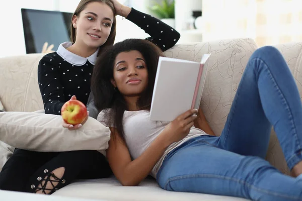 Best friends reading book chilling on sofa, apple for snack, relaxing atmosphere — Photo