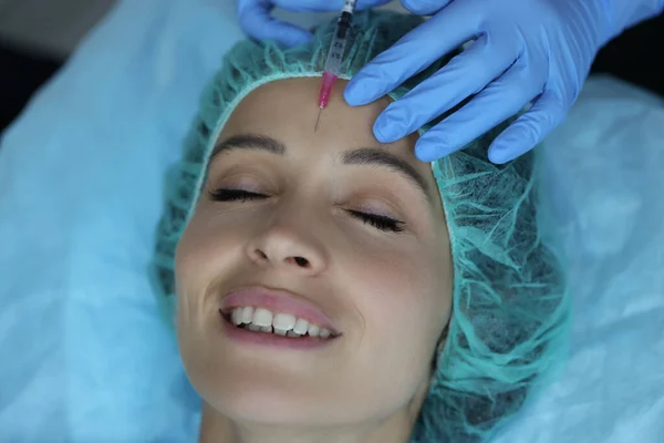 Smiling lady gets injection of botox in her forehead, cosmetologist using syringe with liquid — стоковое фото