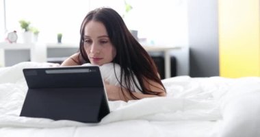 Upset sad woman is working on tablet lying on bed