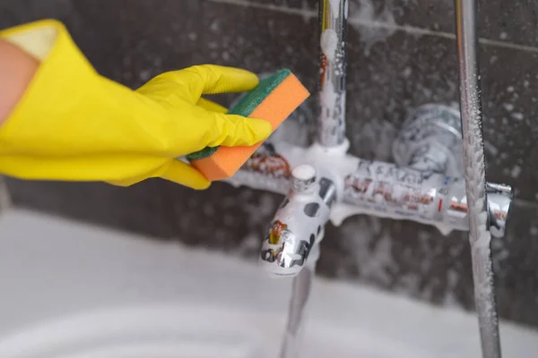 Woman in yellow glove cleans the chrome faucet in bathroom with sponge — Stockfoto