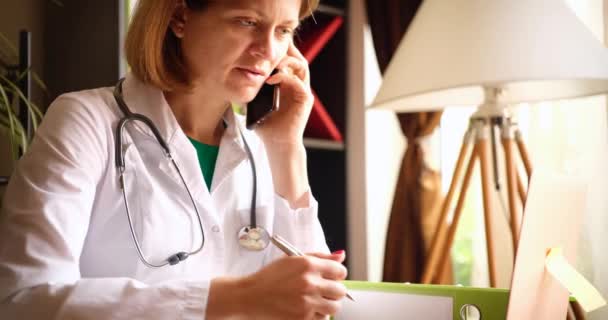 Χαμογελώντας όμορφη γυναίκα γιατρός μιλώντας στο τηλέφωνο στο χώρο εργασίας στην κλινική — Αρχείο Βίντεο