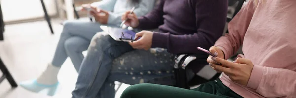 Close-up of people sitting in line and surfing gadgets. Men and women wearing in shoe covers sitting in queue. Waiting for meeting, interview or doctor appointment.