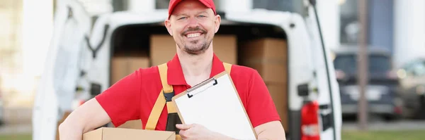 Portrait of happy postman with cardboard package and delivery receipt paper on street. Courier work for big corporation. Private delivery service concept