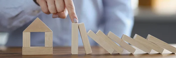 Close-up of female hand stopped falling wooden blocks near home miniature. Property or real estate insurance and security for housing concept. Domino effect idea