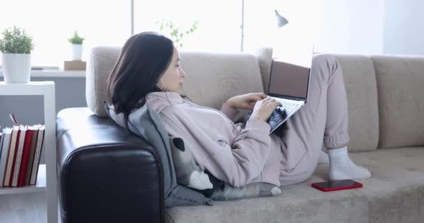 女性がノートパソコンを持つソファの上に家に横たわっています スローモーション リラックスした週末 リモートワークからの休息 — ストック動画