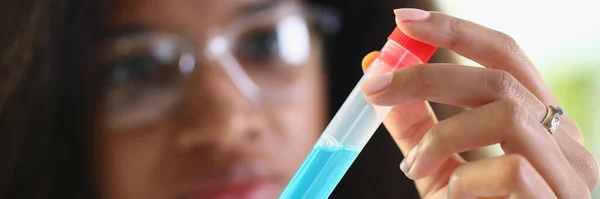 Dark-skinned female laboratory assistant holding a test tube with blue liquid, close-up, blurry. Experiments with drugs for alternative treatment