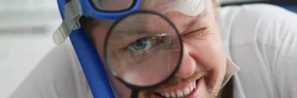 A man wearing a scuba divers mask looks through a magnifying glass, close-up. Search for travel offers, rest from work