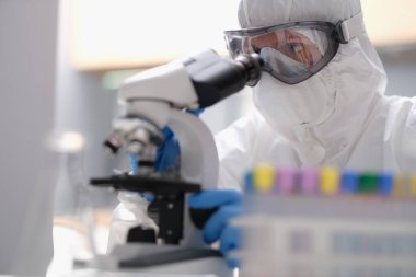 Koruyucu kıyafetli bilim adamı mikroskopla modern laboratuara bakıyor. Bilimsel araştırmalar için yüksek teknoloji kullanarak virüsün evrimini inceleyen laboratuvar asistanı.