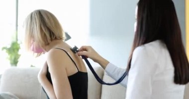 Doktor evde maskeli güçlü öksüren kadının ciğerlerini steteskopla dinliyor. Zatürree ve bronşit kavramı
