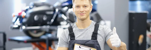 Portrait of young car mechanic holding clipboard and thumb up in car service. Recommendations for high-quality maintenance of cars and motorcycles concept