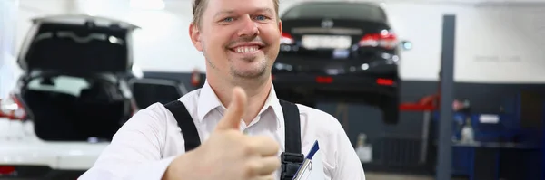 一个笑着的年轻汽车修理工在汽车修理店举起大拇指的画像 优质汽车维修服务概念 — 图库照片